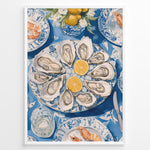 Load image into Gallery viewer, Oyster platter art print plakat showing oysters and lemon slices on a blue patterned table with seafood dishes and glassware in a White frame.
