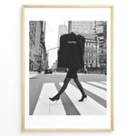 Load image into Gallery viewer, Fashion Plakat | Chic Chanel Street Style Fashion Print Plakaty.dk | Black and white photo of a stylish woman in heels crossing a city street while carrying a Chanel garment bag, framed as wall art.
