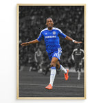 Load image into Gallery viewer, Didier Drogba i Chelsea-trøje med armene strakt ud under en fodboldfejring. Motivet har en sort-hvid stadionbaggrund med fremhævede blå og orange farver.
