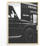 Load image into Gallery viewer, Fashion Plakat | Mercedes-Benz G-Wagon and Prada Store Black and White Art Print Plakaty.dk | A framed, black and white photograph showing the side profile of a Mercedes-Benz G-Wagon AMG parked in front of a Prada store. The car&#39;s V8 BITURBO badge is visible, and the Prada logo is prominent on the building&#39;s facade.

