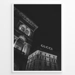 Load image into Gallery viewer, Fashion Plakat | Night View of Gucci Building Facade Plakaty.dk | Black and white nighttime photo of an ornate building with classical columns and arches next to a building displaying a large illuminated &quot;GUCCI&quot; sign against a dark sky.
