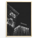 Load image into Gallery viewer, Fashion Plakat | Night View of Gucci Building Facade Plakaty.dk | Black and white nighttime photo of an ornate building with classical columns and arches next to a building displaying a large illuminated &quot;GUCCI&quot; sign against a dark sky.
