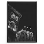 Load image into Gallery viewer, Fashion Plakat | Night View of Gucci Building Facade Plakaty.dk | Black and white nighttime photo of an ornate building with classical columns and arches next to a building displaying a large illuminated &quot;GUCCI&quot; sign against a dark sky.
