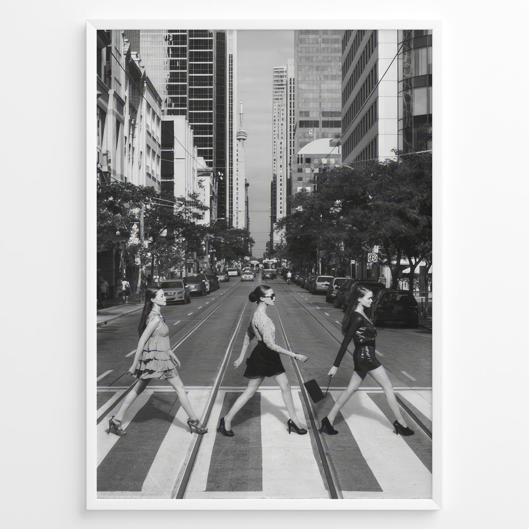Fashion Plakat | Three Women on a Crosswalk in a City Plakaty.dk | Et indrammet sort-hvidt fotografi, der viser tre kvinder, der går fra venstre mod højre på et fodgængerfelt i en by. Kvinden til venstre har en kjole på, kvinden i midten har en top og nederdel, og kvinden til højre har en kort, mørk kjole på og bærer en taske. Høje skyskrabere og et tårn er synlige i baggrunden, med biler parkeret langs gaden.