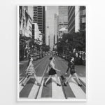 Load image into Gallery viewer, Fashion Plakat | Three Women on a Crosswalk in a City Plakaty.dk | Et indrammet sort-hvidt fotografi, der viser tre kvinder, der går fra venstre mod højre på et fodgængerfelt i en by. Kvinden til venstre har en kjole på, kvinden i midten har en top og nederdel, og kvinden til højre har en kort, mørk kjole på og bærer en taske. Høje skyskrabere og et tårn er synlige i baggrunden, med biler parkeret langs gaden.
