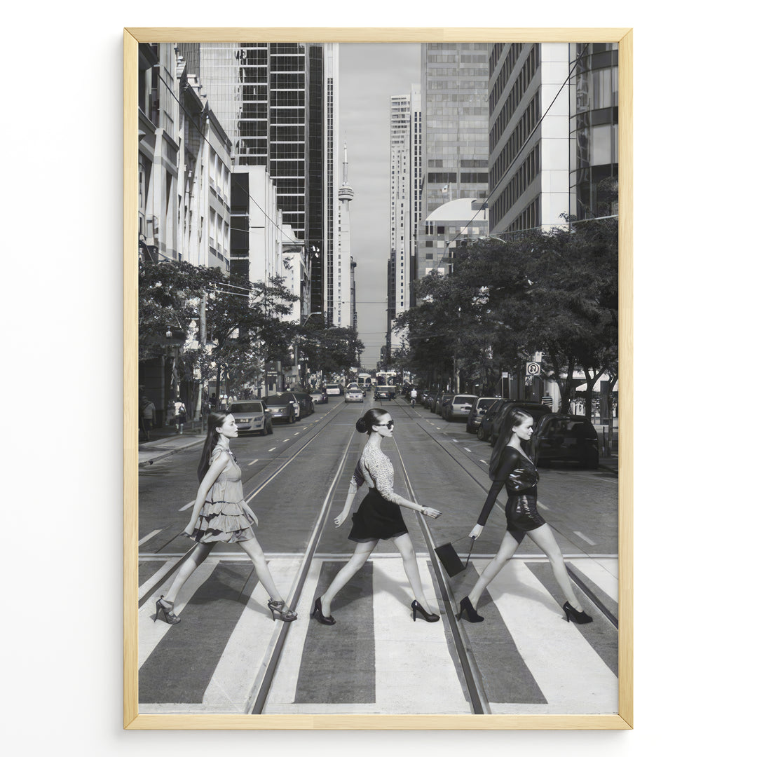 Fashion Plakat | Three Women on a Crosswalk in a City Plakaty.dk | Et indrammet sort-hvidt fotografi, der viser tre kvinder, der går fra venstre mod højre på et fodgængerfelt i en by. Kvinden til venstre har en kjole på, kvinden i midten har en top og nederdel, og kvinden til højre har en kort, mørk kjole på og bærer en taske. Høje skyskrabere og et tårn er synlige i baggrunden, med biler parkeret langs gaden.