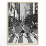 Load image into Gallery viewer, Fashion Plakat | Three Women on a Crosswalk in a City Plakaty.dk | Et indrammet sort-hvidt fotografi, der viser tre kvinder, der går fra venstre mod højre på et fodgængerfelt i en by. Kvinden til venstre har en kjole på, kvinden i midten har en top og nederdel, og kvinden til højre har en kort, mørk kjole på og bærer en taske. Høje skyskrabere og et tårn er synlige i baggrunden, med biler parkeret langs gaden.
