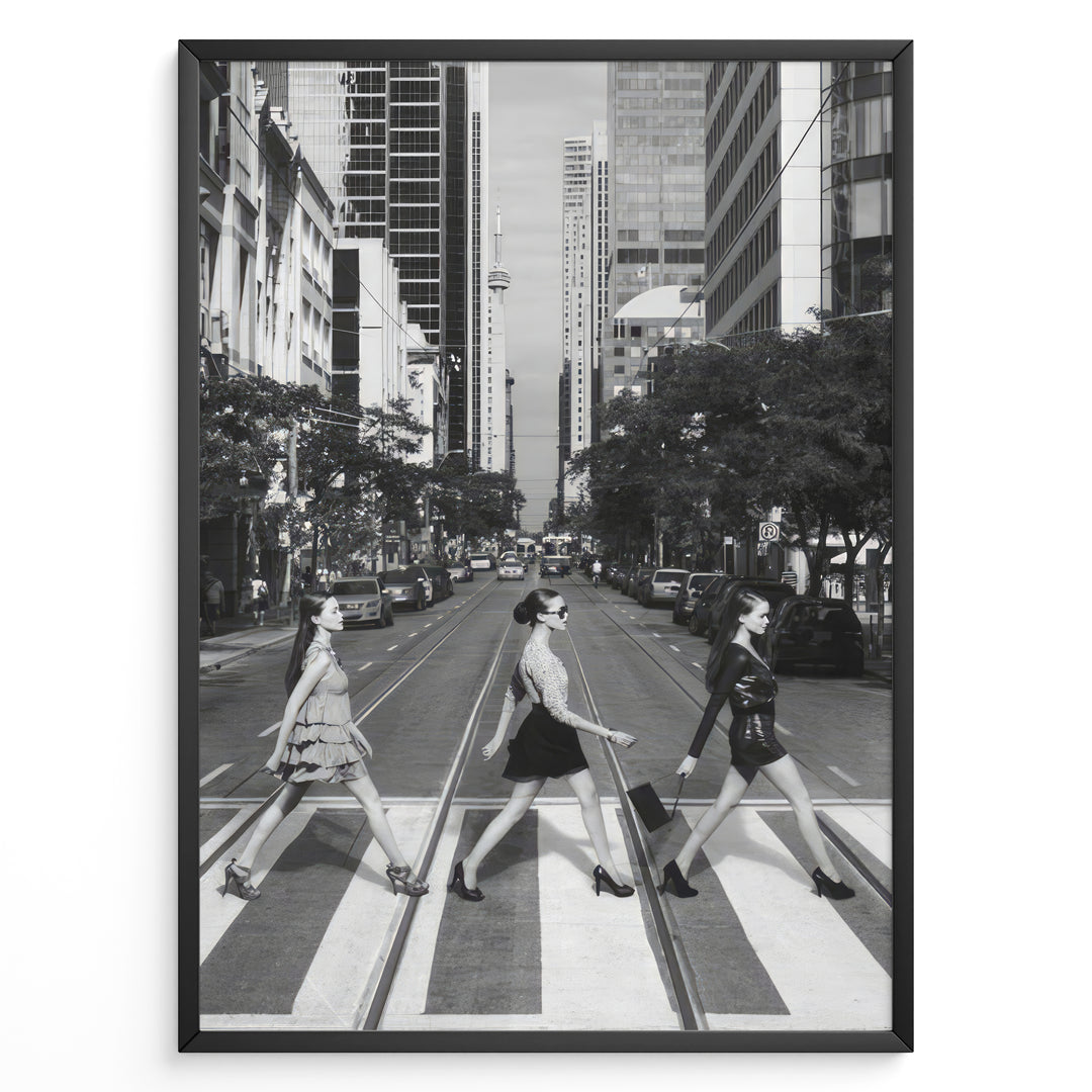Fashion Plakat | Three Women on a Crosswalk in a City Plakaty.dk | Et indrammet sort-hvidt fotografi, der viser tre kvinder, der går fra venstre mod højre på et fodgængerfelt i en by. Kvinden til venstre har en kjole på, kvinden i midten har en top og nederdel, og kvinden til højre har en kort, mørk kjole på og bærer en taske. Høje skyskrabere og et tårn er synlige i baggrunden, med biler parkeret langs gaden.