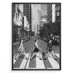 Load image into Gallery viewer, Fashion Plakat | Three Women on a Crosswalk in a City Plakaty.dk | Et indrammet sort-hvidt fotografi, der viser tre kvinder, der går fra venstre mod højre på et fodgængerfelt i en by. Kvinden til venstre har en kjole på, kvinden i midten har en top og nederdel, og kvinden til højre har en kort, mørk kjole på og bærer en taske. Høje skyskrabere og et tårn er synlige i baggrunden, med biler parkeret langs gaden.
