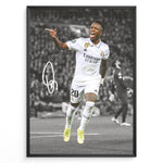 Load image into Gallery viewer, Framed plakat of Vinícius Jr. in a Real Madrid kit celebrating with a pointing gesture, color-highlighted against a black and white background with signature
