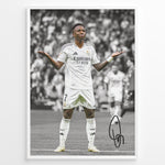 Load image into Gallery viewer, Framed Plakat of Vinícius Jr. in a Real Madrid kit doing a shrug celebration, with a black and white background and visible signature
